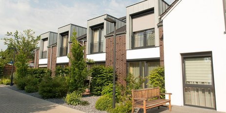 Eine ruhige Wohngegend mit stilvollen, hellen Häusern und einer Holzbank im Vordergrund, umgeben von Pflanzen.