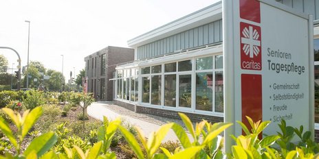 Das Schild der Caritas-Tagespflege für Senioren zeigt wichtige Werte wie Gemeinschaft und Selbstständigkeit, umgeben von einer gepflegten Landschaft.