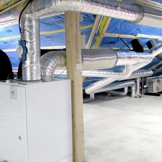 In einem Dachboden sind zahlreiche silberne Lüftungsrohre zu sehen, die sich über eine weiße Heizungsanlage schlängeln.