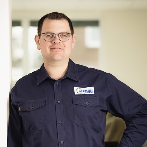 In einem gut beleuchteten Raum posiert ein junger Mann in einem blauen Arbeitshemd mit dem Logo 'TAPKEN'.