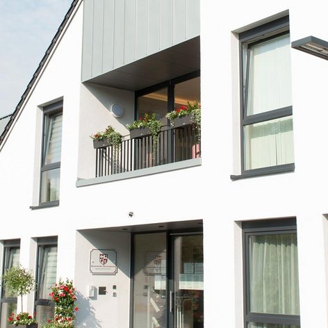 Ein helles, freundliches Wohngebäude mit einem markanten Balkon, der mit blühenden Pflanzen dekoriert ist, und einer einladenden Eingangstür.