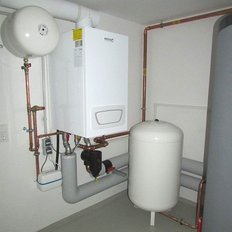 Ein moderner Heizkessel ist an der Wand montiert, umgeben von Kupferrohren und einem großen weißen Druckbehälter.
