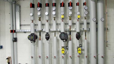 Eine Wand mit mehreren grauen Rohren, Druckmessgeräten und Ventilen, die in einem technischen Raum installiert sind.