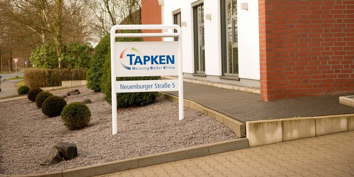 Das Schild von TAPKEN ist gut sichtbar und hebt sich von der grünen Bepflanzung und dem grauen Kies ab.