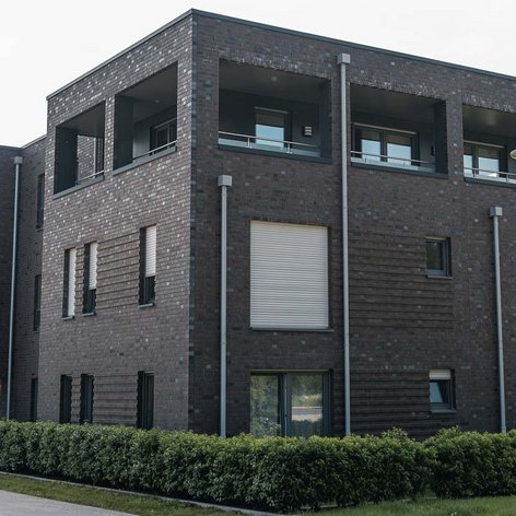 Ein modernes, mehrstöckiges Gebäude mit einer dunklen Ziegelfassade und großen Fenstern, umgeben von gepflegten Sträuchern.