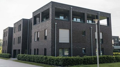 Ein modernes, mehrstöckiges Gebäude mit einer dunklen Ziegelfassade und großen Fenstern, umgeben von gepflegten Sträuchern.