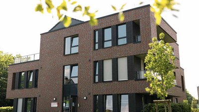 Die Fassade des Hauses zeigt große Fenster und Balkone, die von frischem Grün umgeben sind, was eine einladende Atmosphäre schafft.