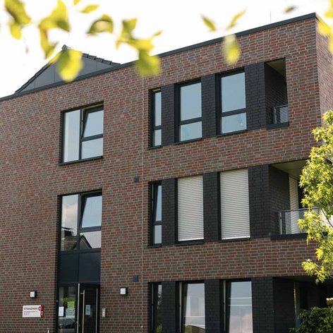 Die Fassade des Hauses zeigt große Fenster und Balkone, die von frischem Grün umgeben sind, was eine einladende Atmosphäre schafft.