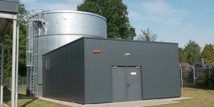 Das Bild zeigt eine moderne Anlage mit einem großen, metallischen Wassertank und einem angrenzenden, grauen Gebäude in der Natur.