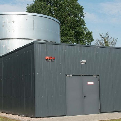 Das Bild zeigt eine moderne Anlage mit einem großen, metallischen Wassertank und einem angrenzenden, grauen Gebäude in der Natur.