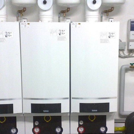 In einem technischen Raum sind drei Heizgeräte installiert, die mit Rohren und einem Steuergerät verbunden sind.