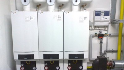In einem technischen Raum sind drei Heizgeräte installiert, die mit Rohren und einem Steuergerät verbunden sind.