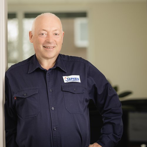 Der Mann mit Glatze trägt ein Hemd mit dem Logo 'TAPKEN' und strahlt Freundlichkeit in einem hellen Raum aus.