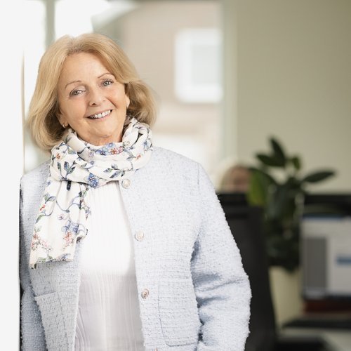 In einem modernen Büro lächelt eine Frau mittleren Alters, die einen blauen Blazer und einen bunten Schal trägt.