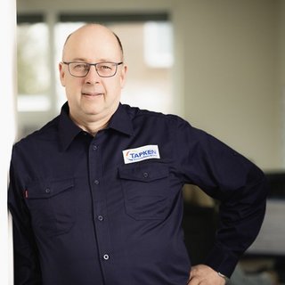 Mann mit Tapken-Logo in entspannter Pose Der Mann mit Brille und einem TAPKEN-Logo auf seinem Hemd steht entspannt an einer Wand in einem modernen Büro.