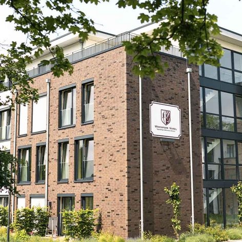 Das Gebäude, umgeben von Bäumen und Sträuchern, zeigt eine einladende Fassade mit einem Schild, das 'Wohnpark Varel' anzeigt.