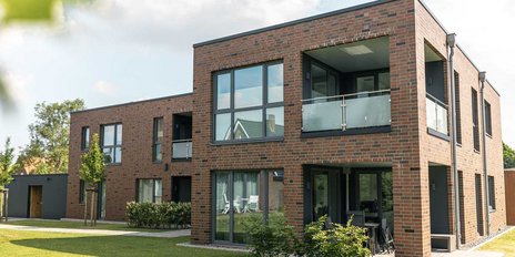 Ein modernes, zweigeschossiges Gebäude aus roten Ziegeln mit großen Fenstern und einem Balkon, umgeben von grünem Rasen.
