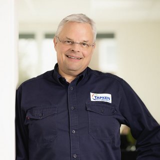 Professioneller Mann in Arbeitskleidung mit Firmenlogo Ein freundlicher Mann mit Brille steht in einem hellen Raum, sein Hemd trägt das Logo eines Unternehmens namens 'TAPKEN'.