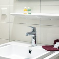 Ein modernes Waschbecken mit einem glänzenden Wasserhahn, umgeben von weißen Fliesen und einem roten Handtuch.