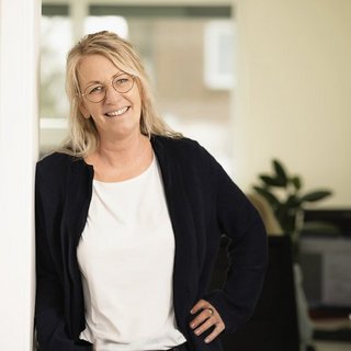 Professionelle Atmosphäre: Frau steht lächelnd im Büro In einem hellen Büro lächelt eine Frau mit Brille, die in einem schwarzen Cardigan gekleidet ist, selbstbewusst in die Kamera.