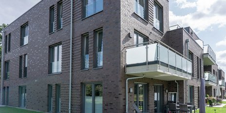 Moderne Wohnanlage mit brauner Ziegelsteinfassade und großen Fenstern, umgeben von grünem Rasen und Bäumen.