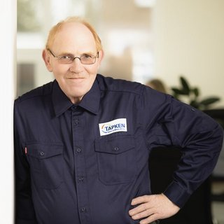 Freundlicher Mitarbeiter in Arbeitskleidung mit Brille Der Mann trägt ein dunkelblaues Hemd mit dem Logo 'TAPKEN' und zeigt eine entspannte Haltung, während er in die Kamera blickt.
