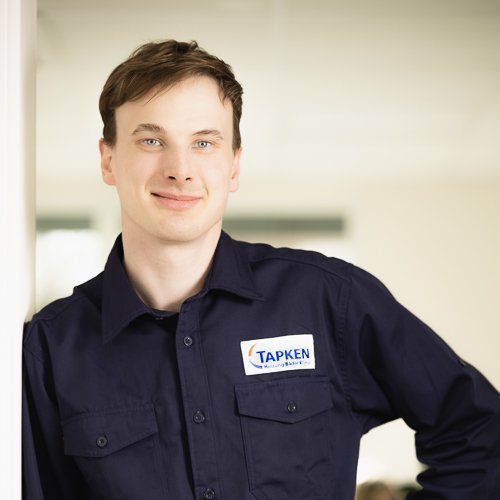 Ein junger Mann mit einem freundlichen Lächeln steht an einer Wand und trägt ein dunkelblaues Hemd mit einem Namensschild.