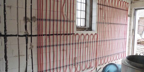 In einem unfertigen Raum sind rote Rohre für die Heizung sichtbar, die an der Wand entlang verlaufen und ein Fenster zeigen.