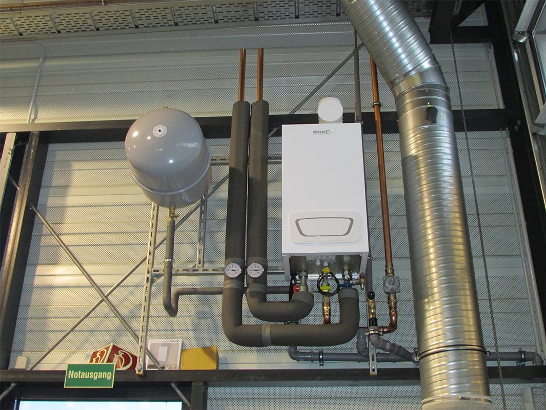 Eine weiße Heizungsanlage mit Rohren und einem Druckmesser, montiert an einer industriellen Wand mit Metallverkleidung.