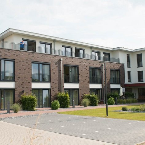 Moderne Wohnanlage mit mehreren Etagen, begrünten Außenbereichen und einem Bewohner auf dem Balkon im Hintergrund.